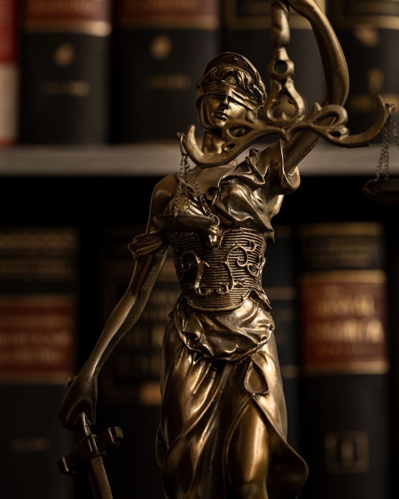 Estátua da Justiça em bronze, de olhos vendados, segurando a balança e a espada diante de livros jurídicos.